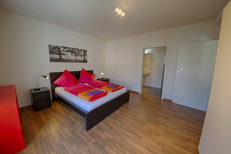 Spacious bedroom with hardwood flooring, white walls, a large black bed frame with colorful bedding, a black nightstand, and a framed artwork on the wall.