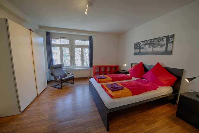 Spacious bedroom with large windows, hardwood flooring, a king-size bed with colorful bedding, a red storage unit, and an armchair. The room has a modern and cozy feel.