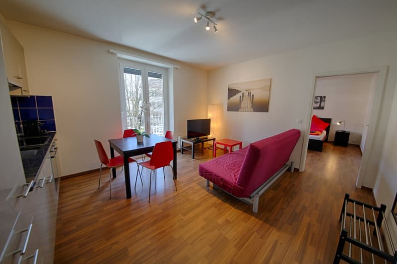 Spacious open-plan living area with hardwood floors, white walls, large windows, dining table with red chairs, red sofa, TV stand, and artwork on the walls.