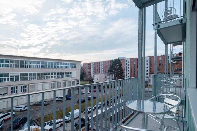 Multi-story apartment building with glass windows, balconies, and a view of the surrounding neighborhood with other residential buildings and parked cars