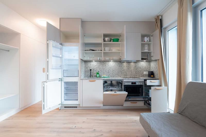 Compact kitchen with white cabinets, granite countertop, built-in appliances including a dishwasher, and open shelving for storage