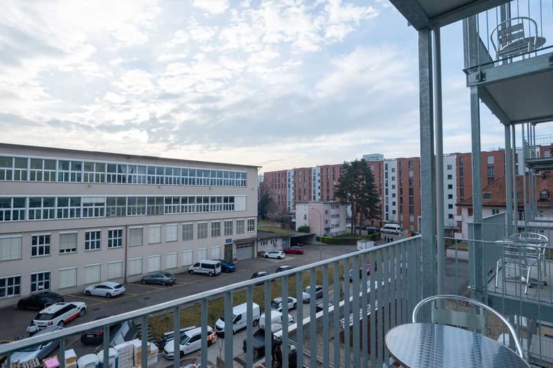 The image shows a multi-story office or apartment building with a balcony or terrace visible. The building has large windows and is surrounded by other residential buildings. There are cars parked in the lot below the balcony.