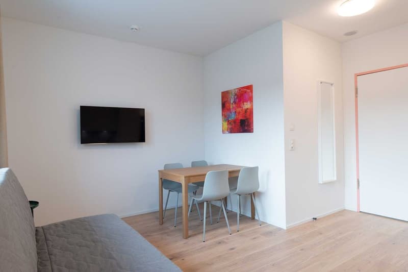 Spacious living room with hardwood floors, white walls, a large TV mounted on the wall, a dining table with four chairs, and a colorful abstract painting on the wall.
