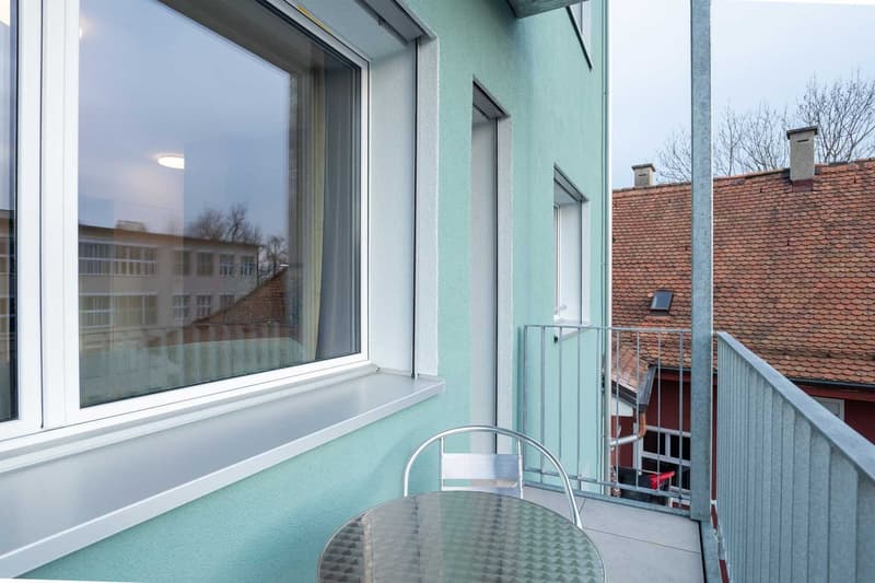 Balcony with glass windows, overlooking a residential building with red tile roof. The balcony has a small table and chair.