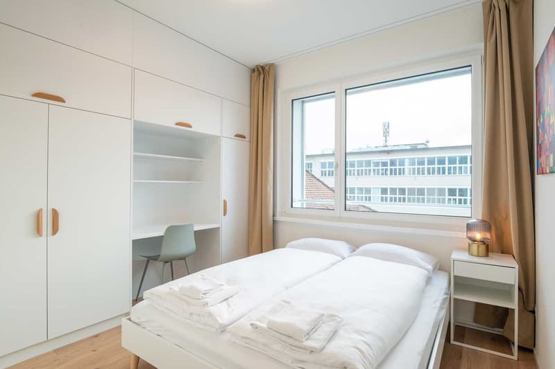 Spacious bedroom with large windows, white walls, wooden flooring, built-in wardrobe, desk, and bedside table with a lamp