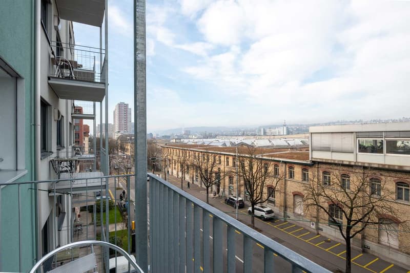 The image shows a balcony or terrace of an apartment building. The balcony overlooks a city skyline with a mix of modern and historic buildings. There are trees and cars visible on the street below.