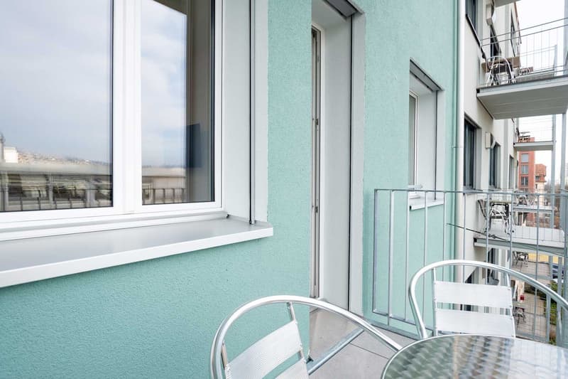 Spacious balcony with large windows, mint green walls, and metal railing. The balcony overlooks a cityscape with other buildings in the background.