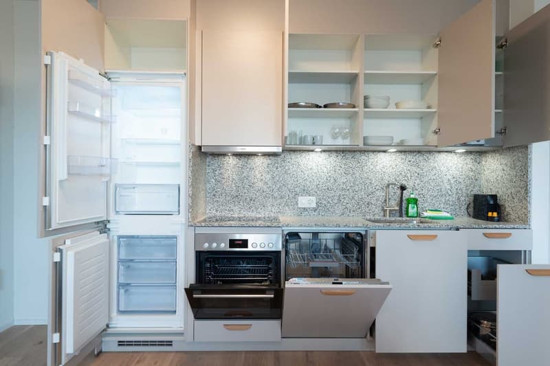 Modern kitchen with white cabinets, granite countertops, built-in oven, dishwasher, and refrigerator. Open shelving displays dishes and glassware.
