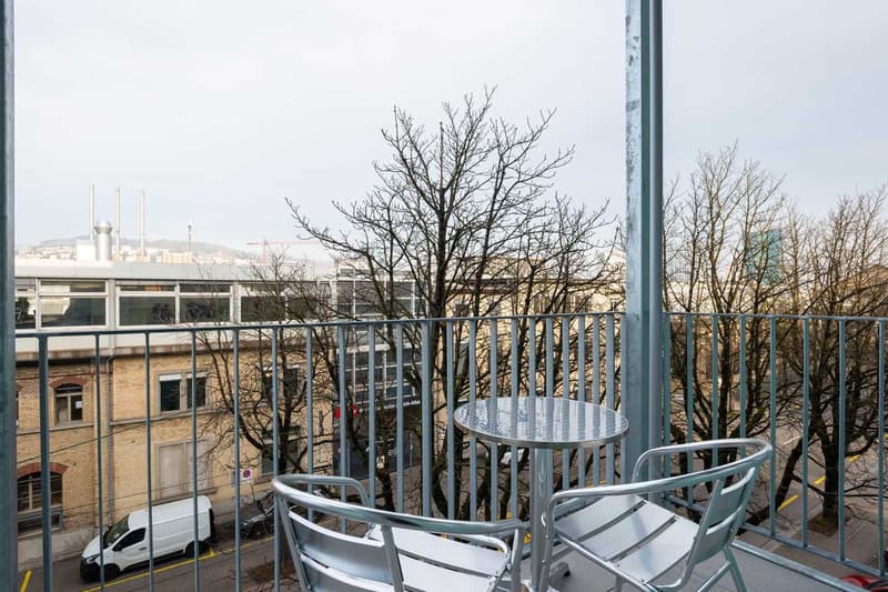 The image shows a balcony or terrace with a metal railing and a small round table with chairs. The view from the balcony includes bare trees and buildings in the distance.
