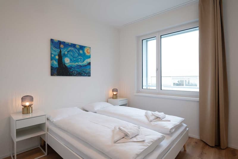 Spacious bedroom with large windows providing a view, white walls, wooden floor, white bedding, nightstands with lamps, and a painting of the Starry Night by Van Gogh on the wall.