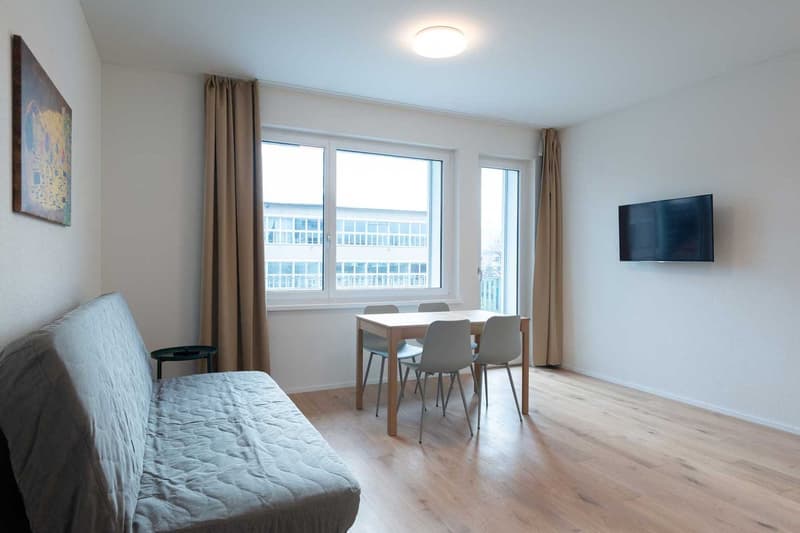 Spacious living room with large windows, hardwood floors, a sofa, a dining table with chairs, and a TV mounted on the wall.