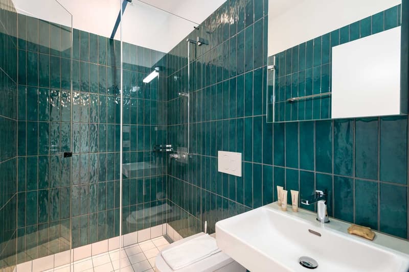Bathroom with teal green tiles covering the walls, a glass shower enclosure, a white sink, and bathroom accessories such as towels and toiletries.