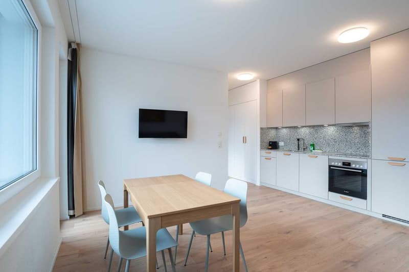 The image shows a modern, open-plan kitchen and dining area. The kitchen features white cabinets, a granite countertop, and a built-in dishwasher. The dining area has a wooden table with four chairs. The room has large windows, providing plenty of natural