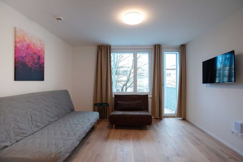 Spacious living room with large windows, hardwood floors, gray sofa, brown leather chair, and a colorful abstract painting on the wall. The room also has access to a balcony or terrace.