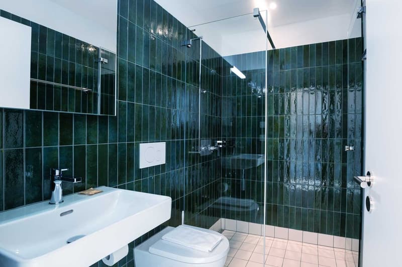 Spacious bathroom with dark green tiled walls, a white sink, a toilet, and a glass-enclosed shower area.