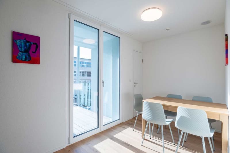 Bright and spacious dining area with large sliding glass doors leading to a balcony or terrace. The room features a wooden dining table with white chairs, and a colorful abstract painting on the wall.