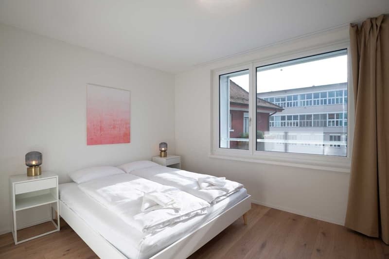 Spacious bedroom with large windows providing a view, white walls, hardwood floors, a double bed with white bedding, and minimalist decor including a framed artwork and bedside lamps.