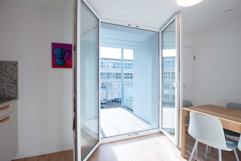 Bright and spacious apartment with large glass sliding doors leading to a balcony or terrace. The room features white walls, hardwood floors, and modern furniture including a dining table and chairs. A colorful abstract painting adds a touch of personalit