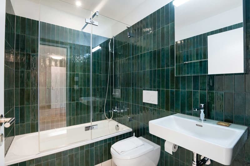 Spacious bathroom with green tiled walls, a glass shower enclosure, a white sink, and a toilet. The bathroom appears modern and well-designed.