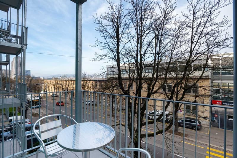 Balcony with a small table and chairs, overlooking a street with bare trees and buildings in the background