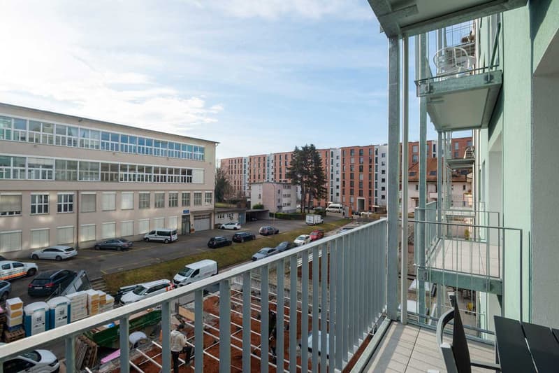 The image shows a multi-story apartment building with a balcony or terrace visible. The building has a modern, glass-heavy design with many windows. There is a parking lot in the foreground with several cars parked. The surrounding area appears to be a re