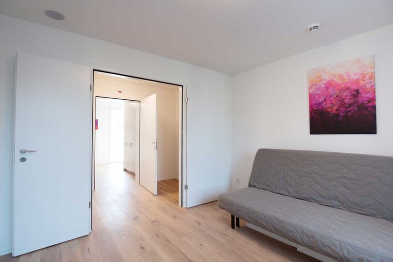 Spacious bedroom with hardwood floors, white walls, large windows, and a colorful abstract painting on the wall. The room features a grey sofa bed and an open doorway leading to another room.