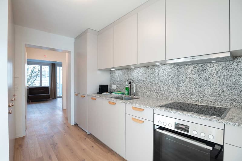Modern kitchen with white cabinets, granite countertops, and stainless steel appliances including a dishwasher. The kitchen leads to an adjacent room through an open doorway.
