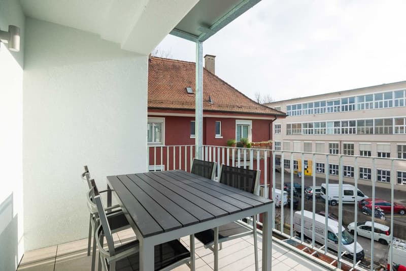The image shows a balcony or terrace area with a large outdoor dining table and chairs. The balcony overlooks a parking lot and other buildings. The building itself has a red-tiled roof and white walls.