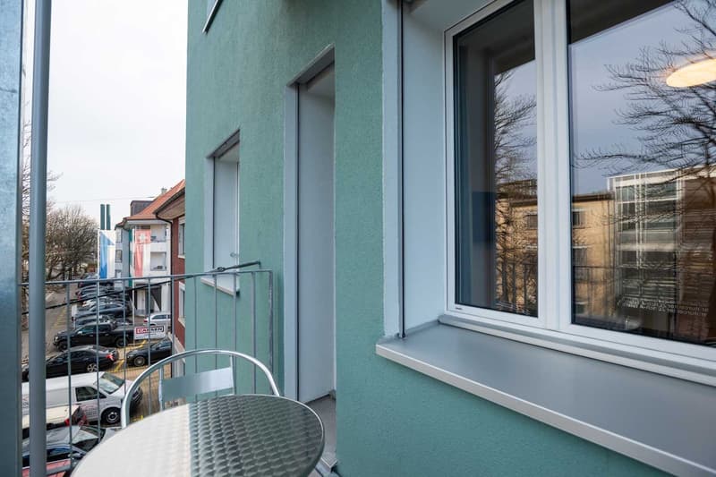 The image shows the exterior of a multi-story building with a green facade. The building has large windows and a balcony or terrace on the upper floor. There are cars parked in front of the building, and the surrounding area appears to be a residential or