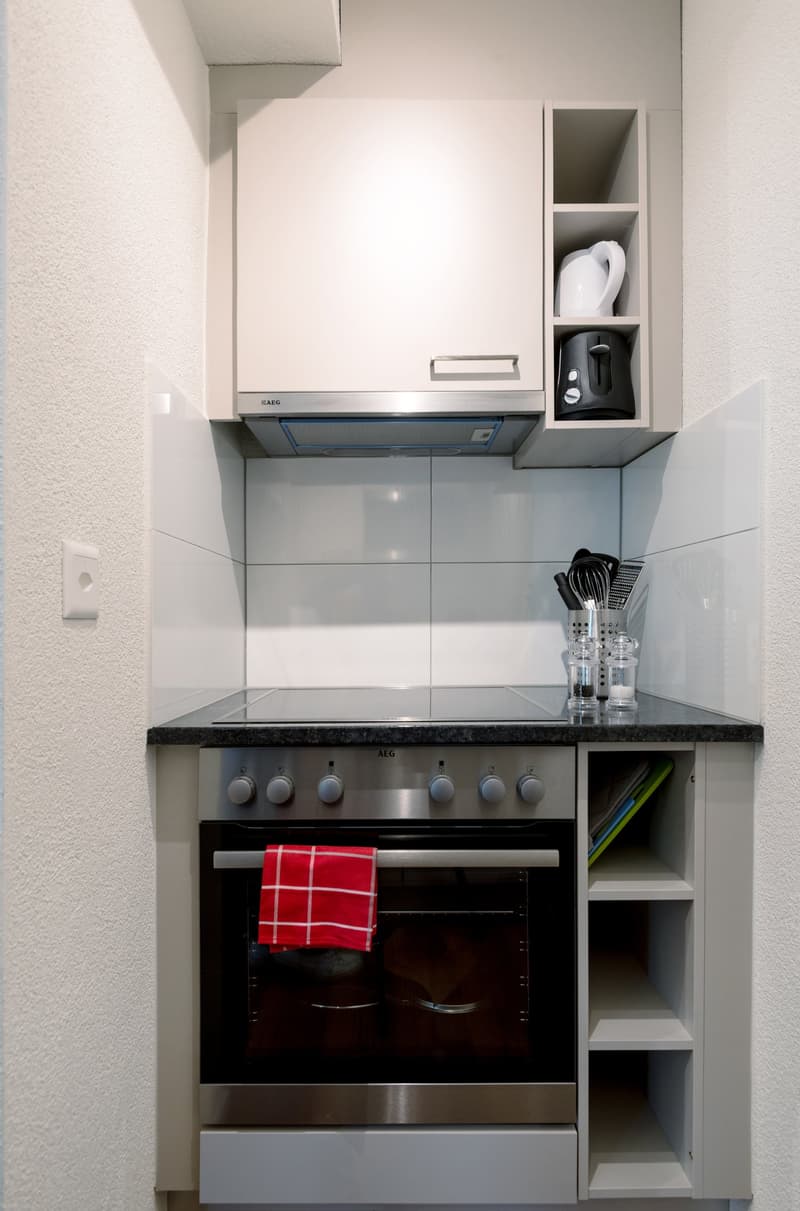 Modern kitchen with white cabinets, black granite countertop, built-in AEG oven and stove, and storage shelves above the oven. A red and white kitchen towel is visible in the oven.