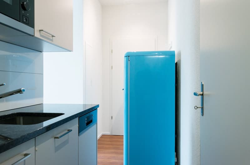 Modern kitchen with blue appliances, including a blue refrigerator and built-in dishwasher, along with a black countertop and white cabinets.