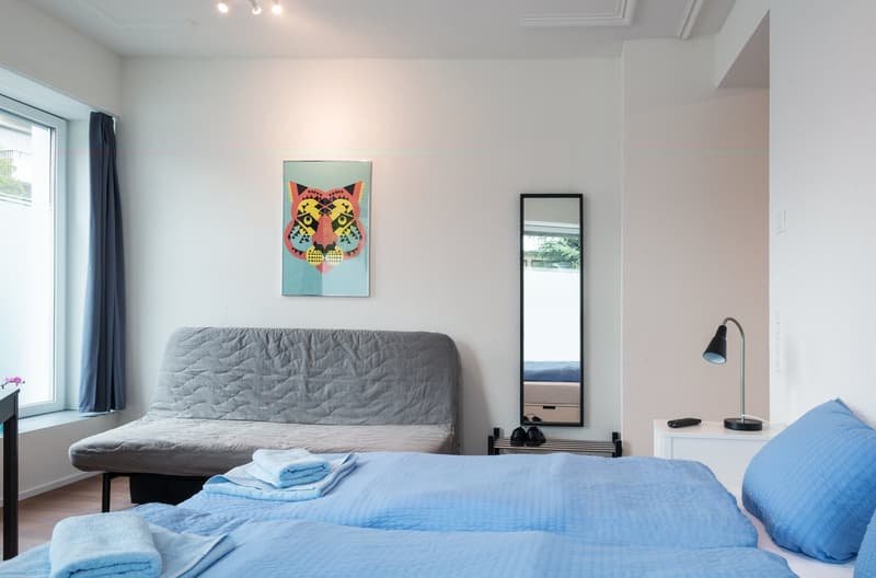 Spacious bedroom with large windows, gray sofa, colorful wall art, and a full-length mirror. The room has a cozy and modern feel with blue bedding and a lamp on the side table.