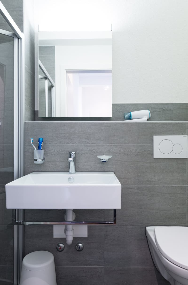 Modern bathroom with white sink, gray tile walls, mirror, and toilet