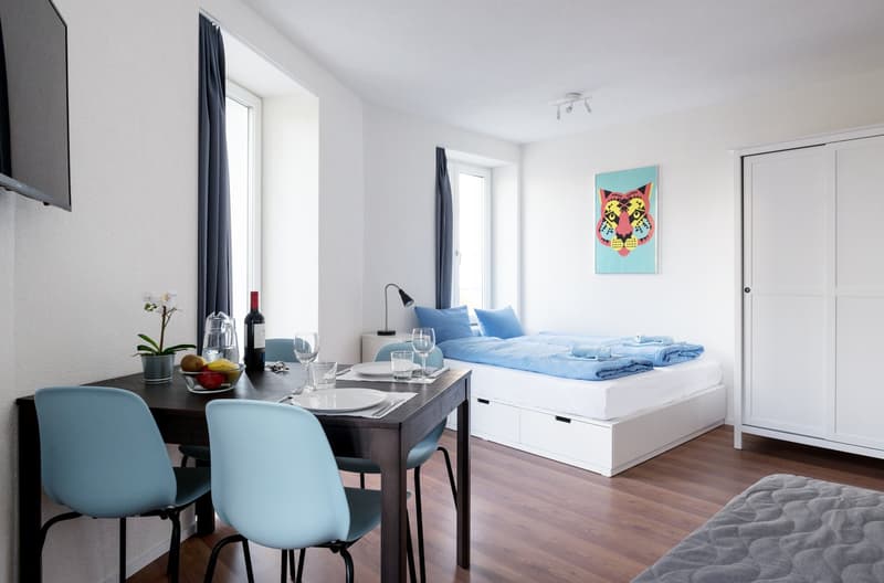 The image shows a studio apartment with an open floor plan. The living and sleeping areas are combined, with a bed and a dining table with chairs. The room has large windows, hardwood floors, and a modern, minimalist decor. There is a TV mounted on the wa