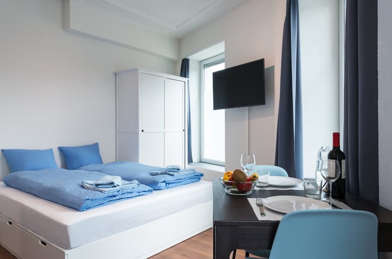 Spacious bedroom with a large bed, blue bedding, and a TV mounted on the wall. There is a wardrobe and a table with a dining setup, including plates, glasses, and a bottle of wine. The room has large windows, providing natural light.