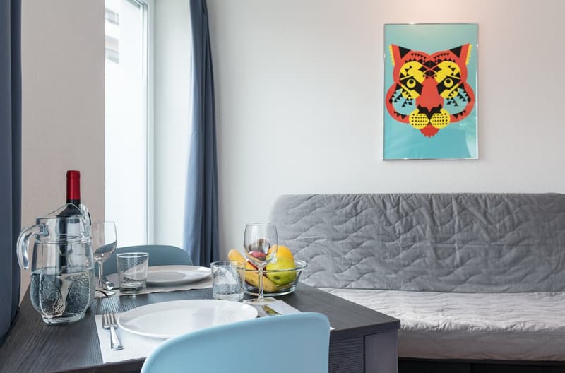 Gray sofa, coffee table with plates and glasses, colorful artwork on the wall, curtains, and a view through the window