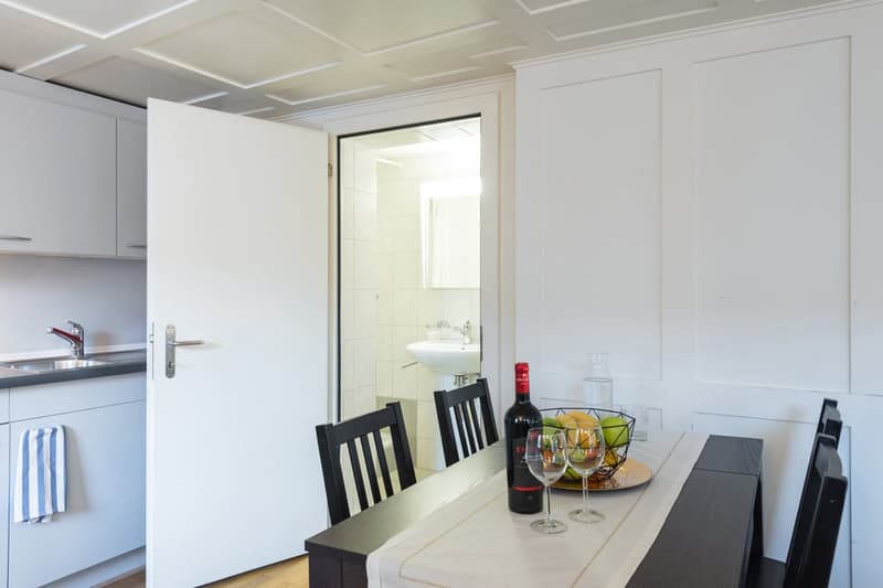 The image shows a kitchen and dining area with white walls, a coffered ceiling, and a dining table with chairs. There is a kitchen sink and cabinets visible, as well as a dishwasher. The room also has a bathroom visible through an open doorway.