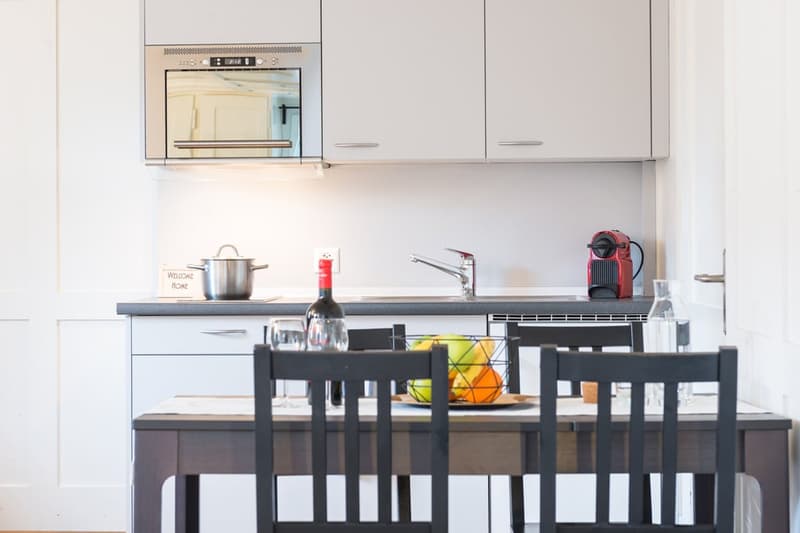 Modern kitchen with white cabinets, black countertop, dining table with chairs, kitchen appliances including a dishwasher, and various kitchen items such as a pot, wine bottle, and fruit