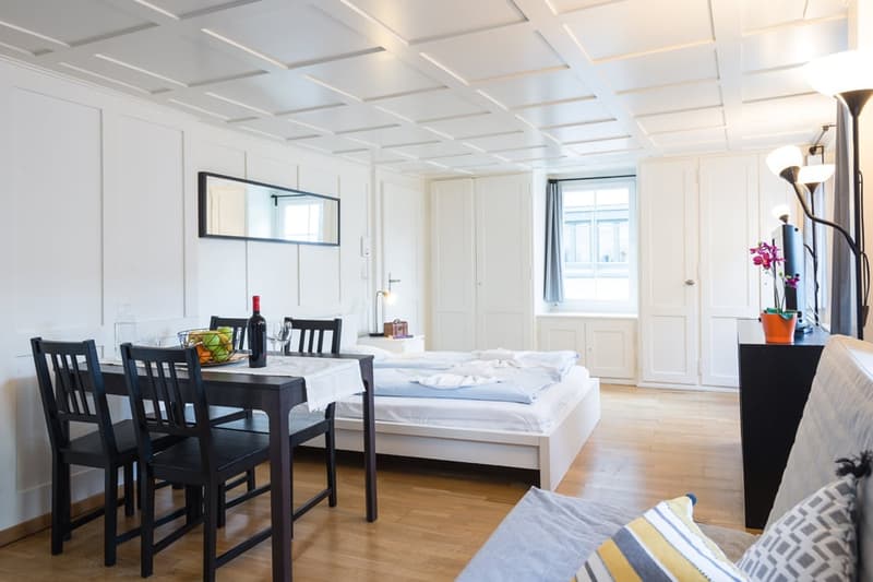 Open-plan studio apartment with a dining area, a bed, and a living room. The room has white walls, a coffered ceiling, and hardwood floors. There is a large window that provides natural light. The furniture includes a dark wooden dining table with chairs,