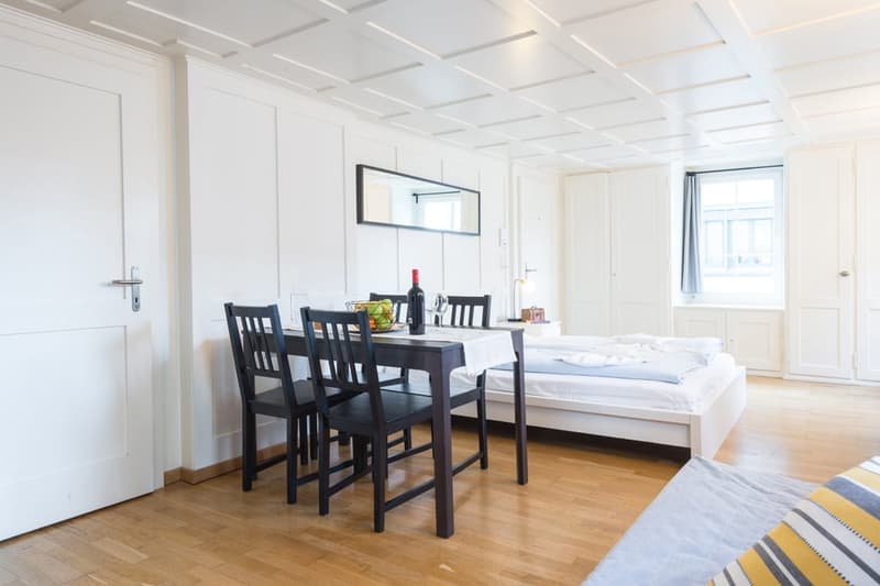 Open-plan studio apartment with dining area, kitchenette, and sleeping area. White walls, hardwood floors, coffered ceiling, black dining table and chairs, bed, and window.