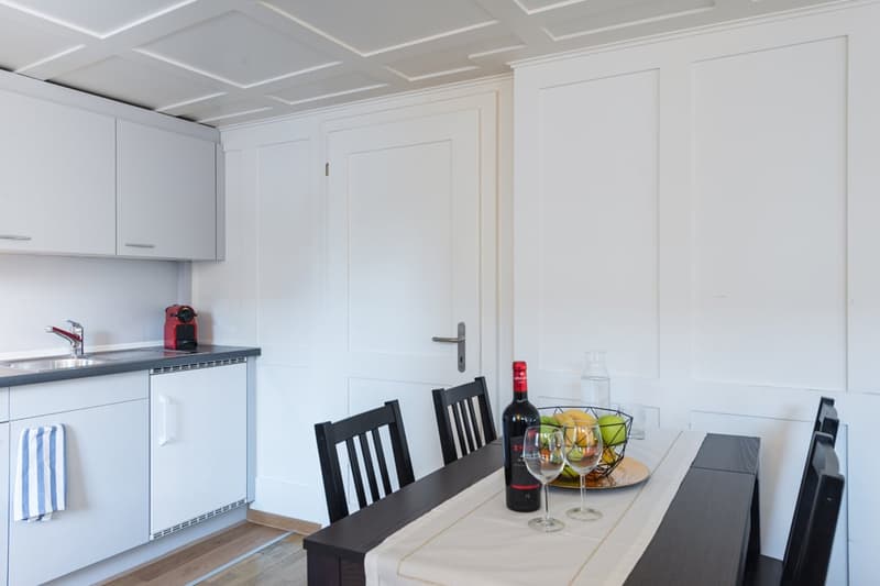 The image shows a modern, open-plan kitchen and dining area. The kitchen features white cabinets, a stainless steel sink, and a dishwasher. The dining area has a wooden table with black chairs, and a basket of fruit on the table. The room has a clean, min