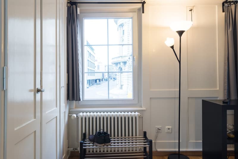This image shows a room with a large window overlooking a city street. The room has white walls, a wooden bench, and a floor lamp. There is a closed door visible on the left side of the image.