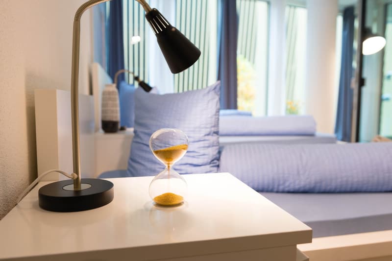 Bedroom with large window, blue curtains, white bedding, and a desk with a lamp and hourglass on it.