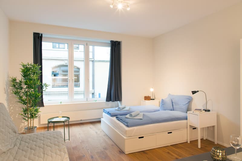 Spacious bedroom with large windows, hardwood floors, white walls, a bed with storage drawers, bedside tables, a potted plant, and a cozy seating area by the window.