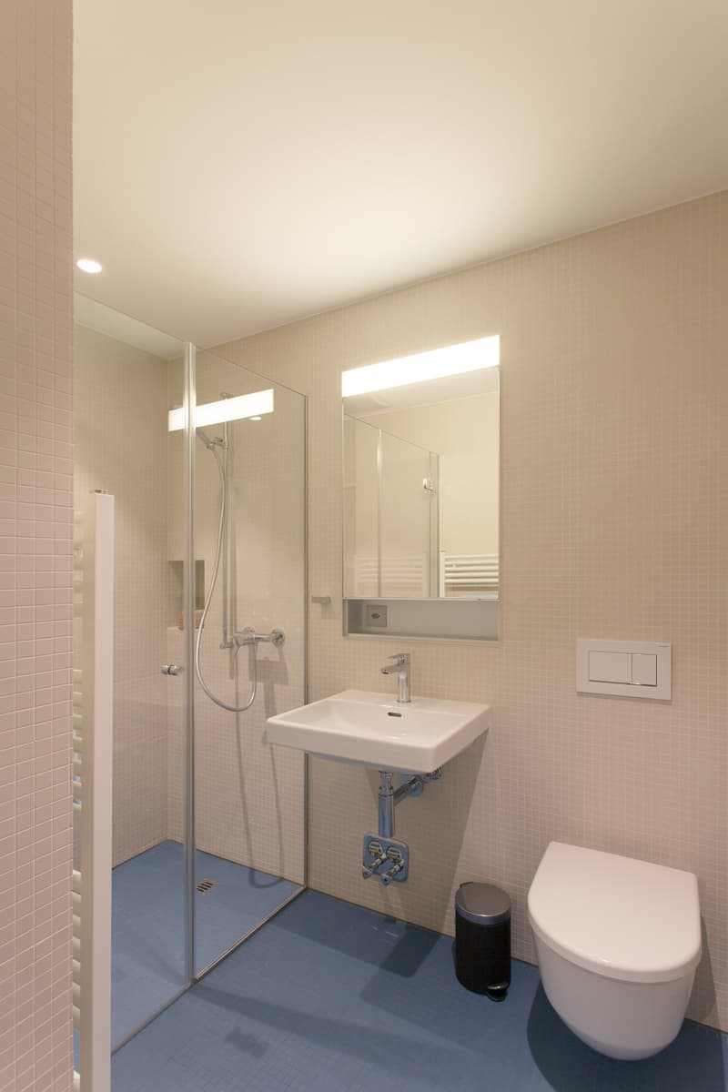 The image shows a modern bathroom with a glass shower enclosure, a white sink vanity, and a toilet. The walls are tiled in a light beige color, and the floor is covered in a blue tile. The bathroom appears to be well-lit with recessed lighting and a mirro