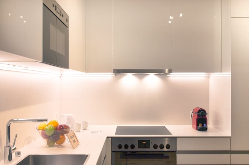 Modern kitchen with white cabinets, built-in oven, induction cooktop, stainless steel sink, and a bowl of fresh fruits on the counter. A Nespresso coffee machine is also visible.