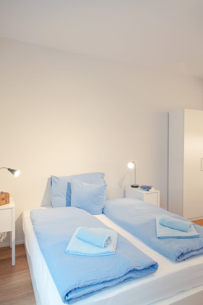 Bedroom with a white bed, blue bedding, and a side table with a lamp. The room has a minimalist and clean design.