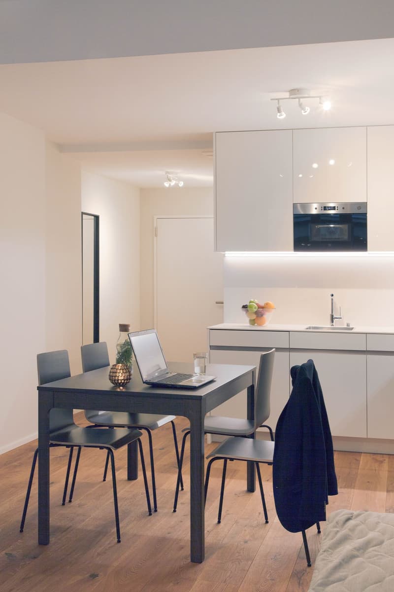 The image shows a modern, open-plan kitchen and dining area. The kitchen features white cabinets and a built-in dishwasher. The dining table is a simple black rectangular table with four chairs. There is a laptop and some decor items on the table, suggest