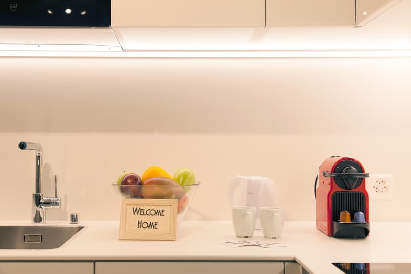 Modern kitchen with stainless steel sink, coffee machine, and a bowl of fresh fruits on the counter. A 'Welcome Home' sign is also visible.