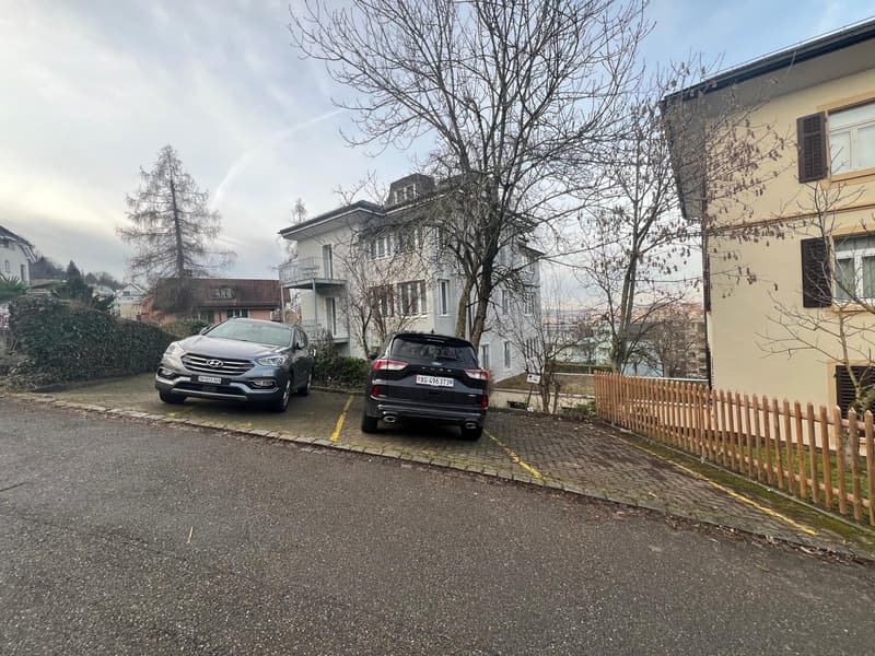The image shows a residential neighborhood with several multi-story buildings, trees, and parked cars on the street. The buildings have a mix of architectural styles, with some appearing older and others more modern. The street is paved with a cobblestone